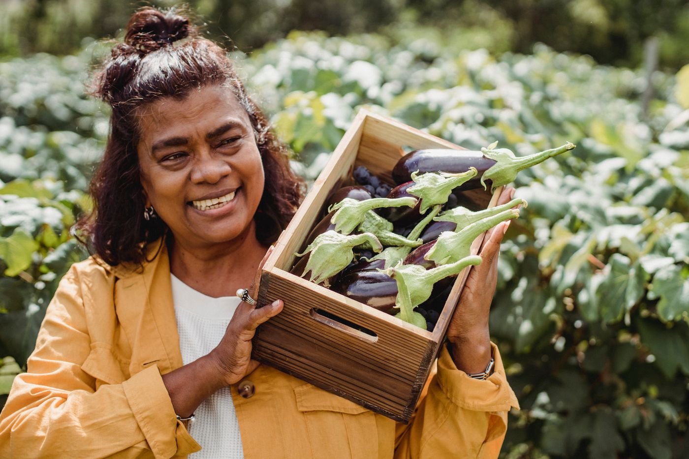 A smiling woman in a yellow jacket holding a wooden crate of freshly picked eggplants